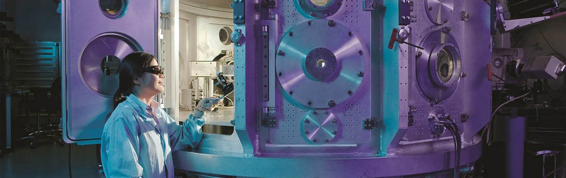 a female researcher sitting with her microscope by an enormous machine